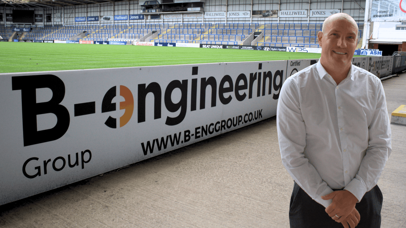 A man in a white sport stand with his hand folded in front of a stadium ground with a banner in the background in white. The banner has the brand name B-engineering