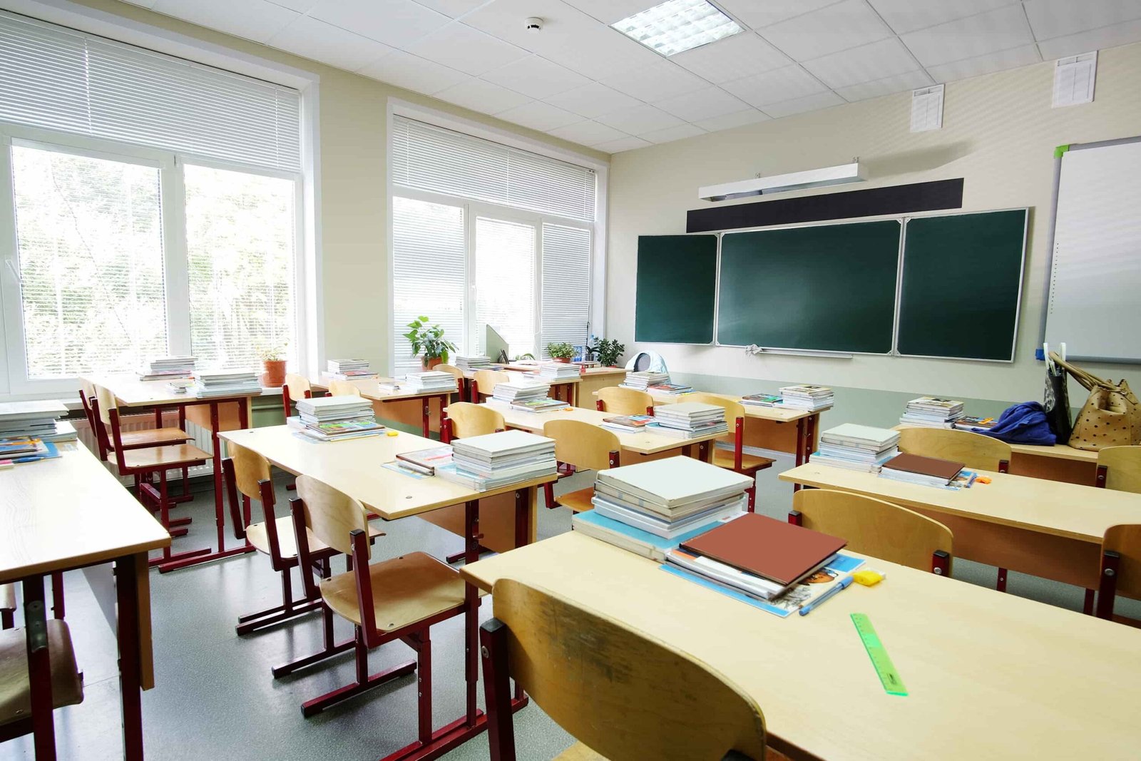 Empty classroom with wooden desks and red chairs, stacks of books, green chalkboard, and large windows. Bright, orderly setting.