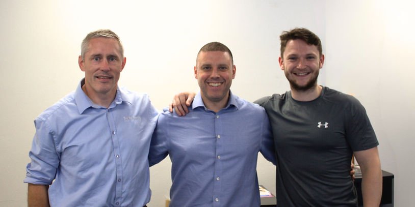 Three men smiling and standing with arms around each other in a white-walled room. Two wear blue shirts, one in a gray T-shirt. Casual, happy mood.
