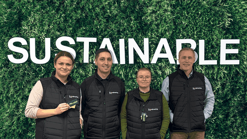 Four people in black vests stand smiling in front of a leafy wall with the word "SUSTAINABLE" in white. Two hold cards.