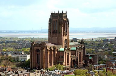 Grand Gothic cathedral towers over a cityscape with green rooftops and distant hills. Bright sky and river in the background.