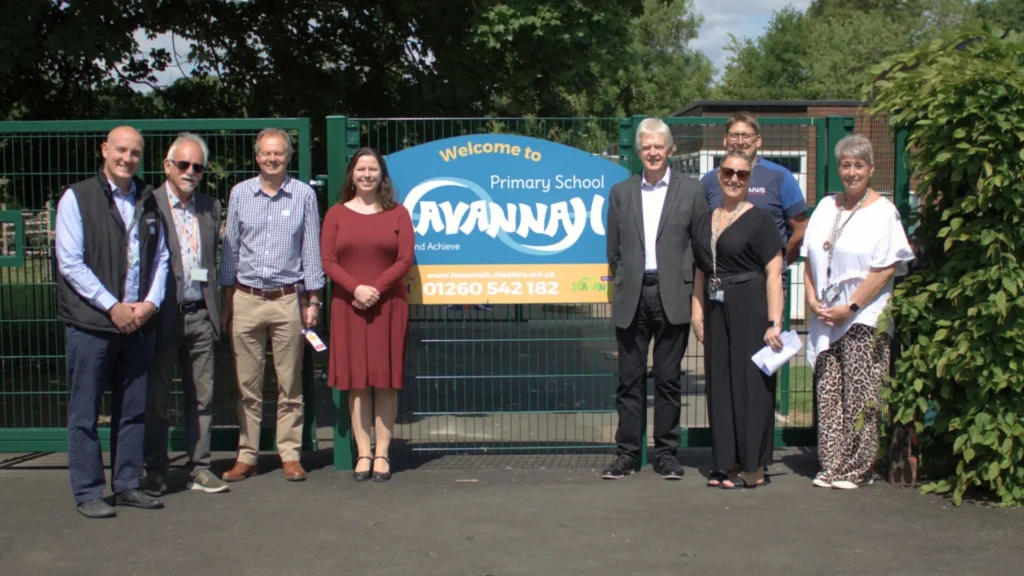 Eight people stand smiling in front of a "Welcome to Havannah Primary School" sign. Sunny day, greenery in the background.
