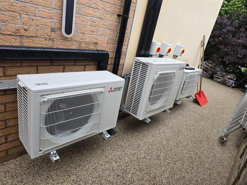 Three Mitsubishi air conditioning units mounted on a brick wall, with a red shovel nearby. Pebbled ground and some foliage in the background.