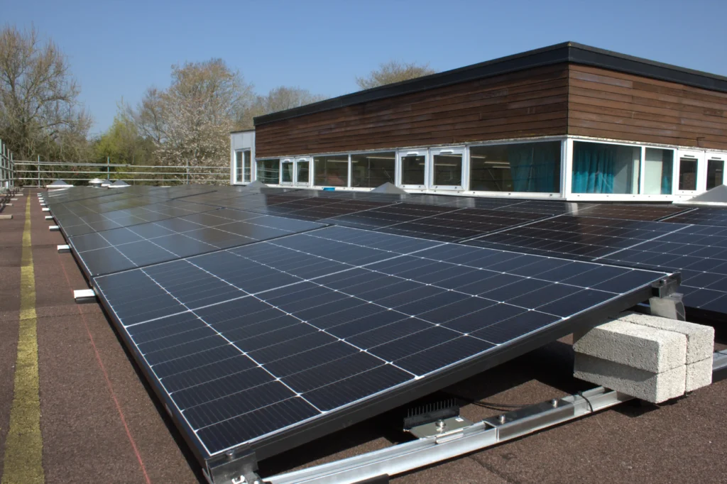 Solar panels on a flat rooftop in a sunny outdoor setting, surrounded by trees. Building with wood paneling and windows in the background.