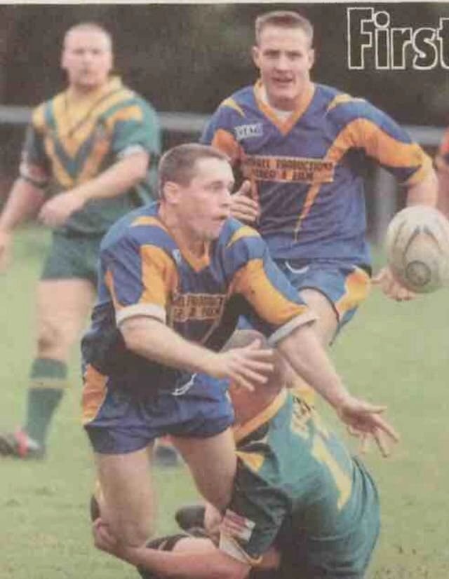 Rugby players in blue and yellow jerseys in action on a grassy field. One player is tackled, passing the ball. Energetic and competitive scene.