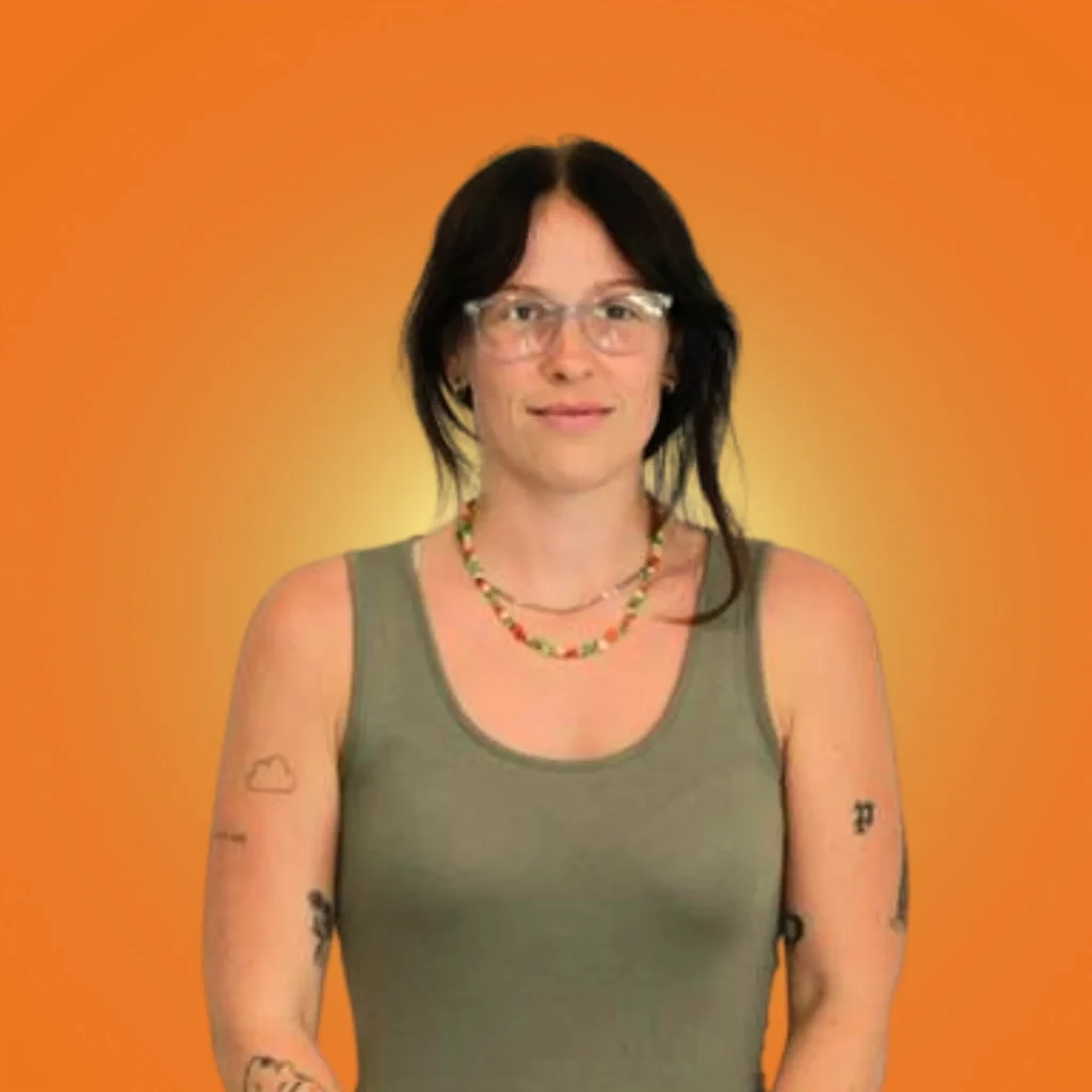 Person with glasses, wearing a green tank top and colorful necklace, stands against an orange background. Tattoos visible; calm mood.
