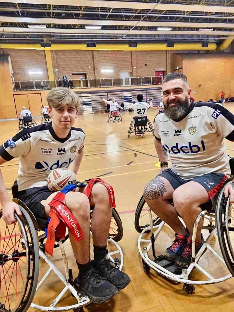Two wheelchair rugby players in white jerseys, holding a rugby ball, smile on an indoor court, with others and brick walls in the background.