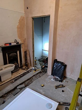 Room under renovation, tools scattered on the floor, a person working near a wall. Exposed pipes and a fireplace visible. Neutral colors.