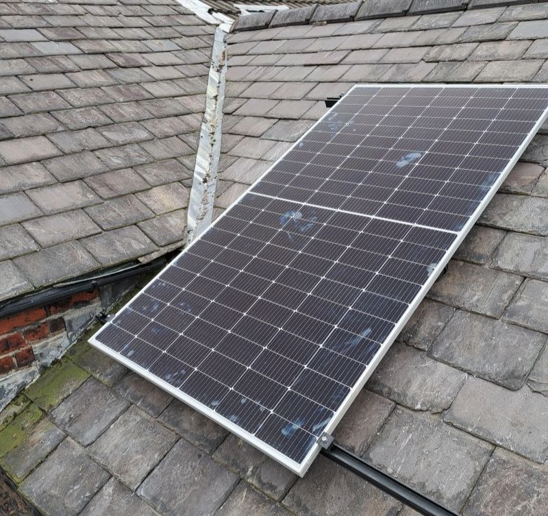 A solar panel installed on a grey tiled roof, with weathered brick and moss visible. Overcast lighting and subtle blue markings on panel.
