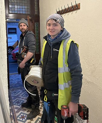 Two workers in winter gear stand in a hallway with patterned tiles. One holds cable and a tool, the other carries a tool belt. Mood is focused.