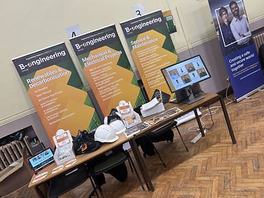Exhibition setup with banners for engineering services, a table with hard hats, monitor, and leaflets. Text reads: Renewables & Decarbonisation.