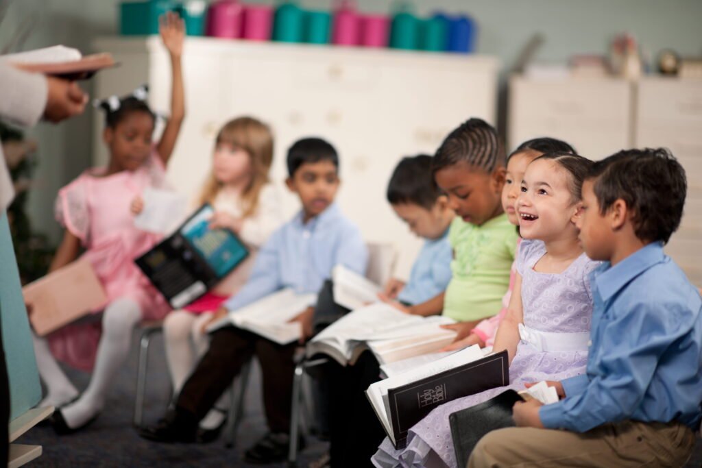 Children sit in a classroom, smiling and holding books, one raising a hand. Shelves with colorful bins are in the background.