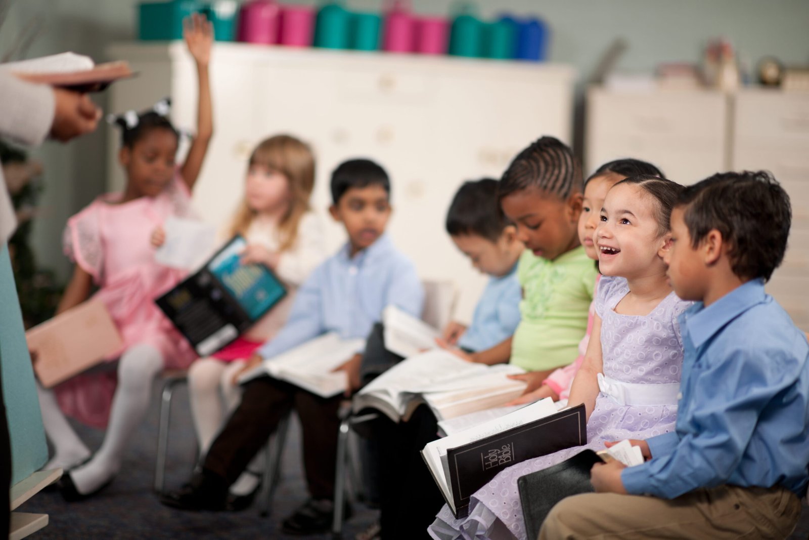 Children sit in a classroom, smiling and holding books, one raising a hand. Shelves with colorful bins are in the background.