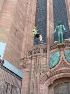 A man dangles from a tall building in a harnest, abseiling down. He wears an orange helmet and green t-shirt.