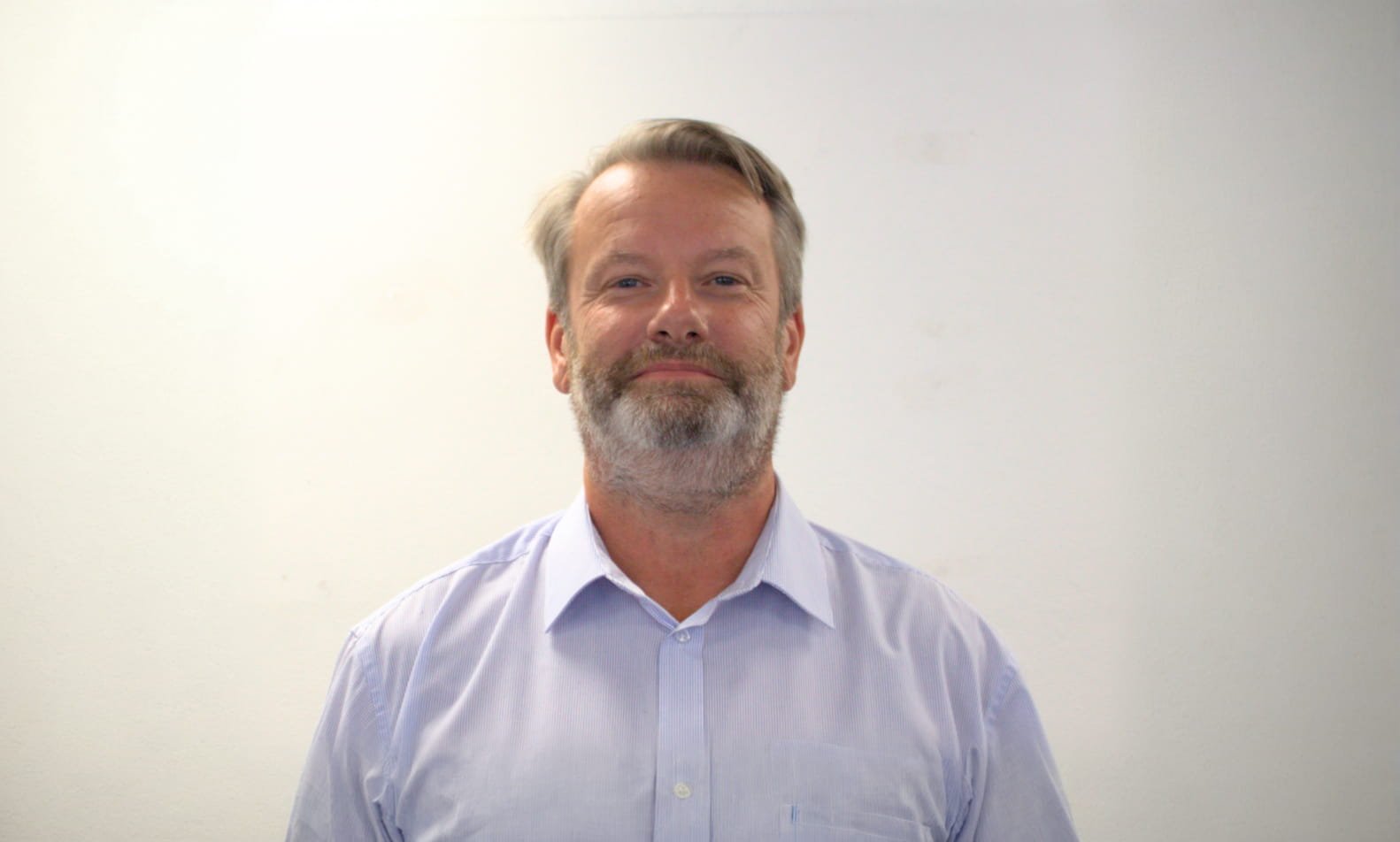 A man in a blue shirt stands in front of a white wall. He has a gray beard anad matching hair
