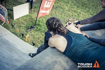 A woman lies on her front at the edge of a cliff drop holding onto a person trying to climb upwards. There are others to the right who hold onto his right arm. 
