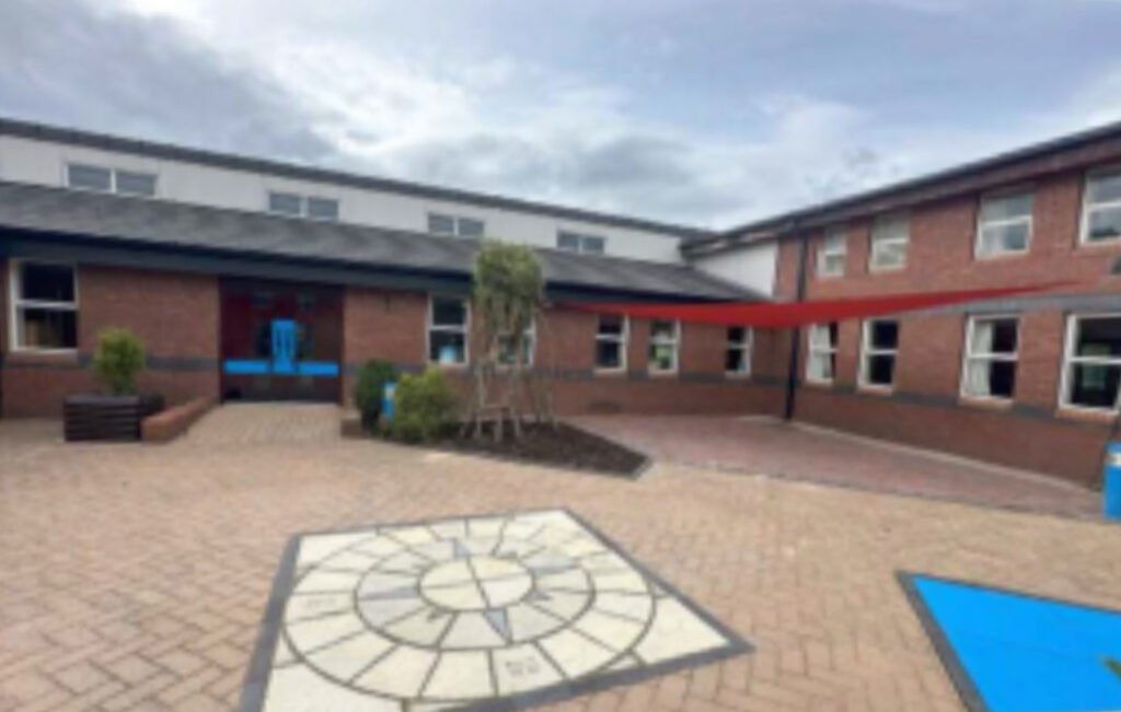 Brick courtyard with a red canopy, geometric pattern on the ground, and plants. Buildings surround the open space under a cloudy sky.