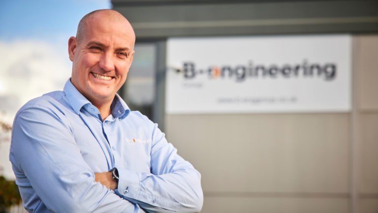 Director of B-engineering, Rob Ditchfield stand in front of company facilities with large branding.