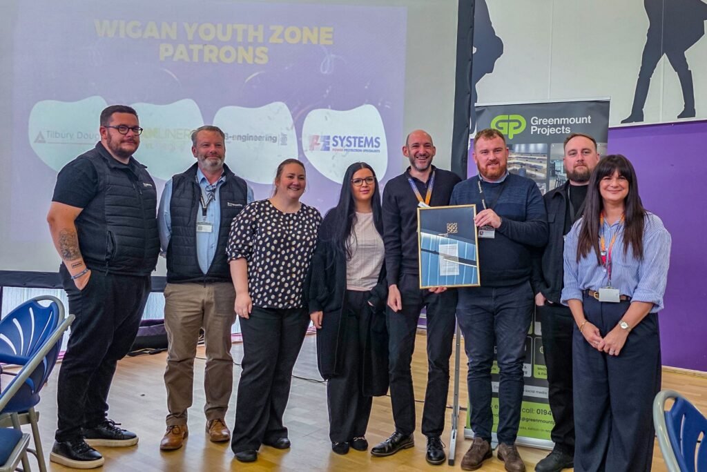 Eight people stand smiling indoors, with "Wigan Youth Zone Patrons" projected behind. One person holds a framed certificate.