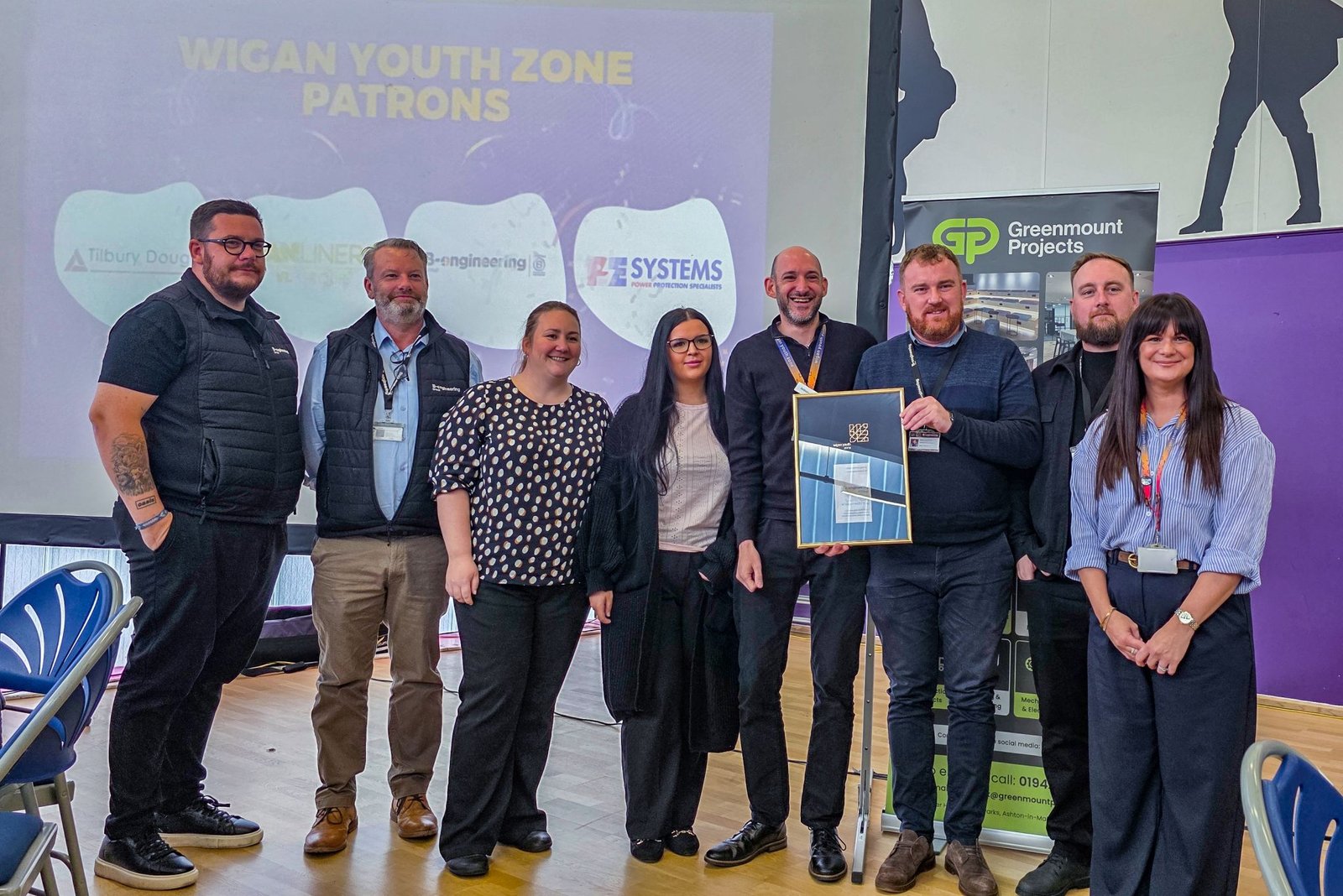 Eight people stand smiling indoors, with "Wigan Youth Zone Patrons" projected behind. One person holds a framed certificate.