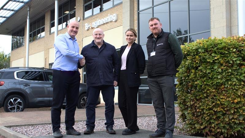 Four people stand smiling outside a building with "Engineering" text. Two men shake hands. A car and bushes are visible in the background