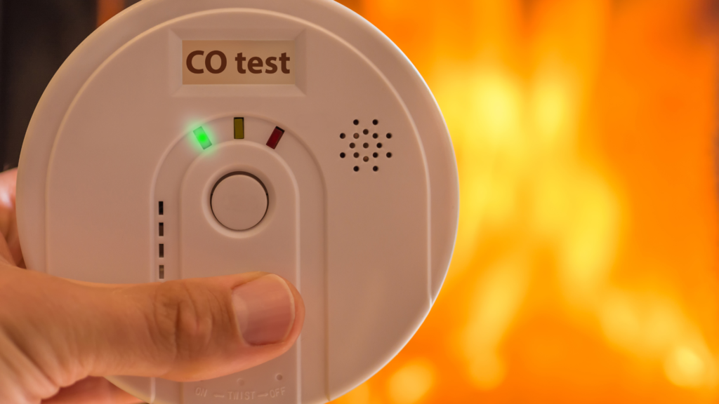 Hand holding a CO detector showing a green light, with flames in the background. The device label reads "CO test."