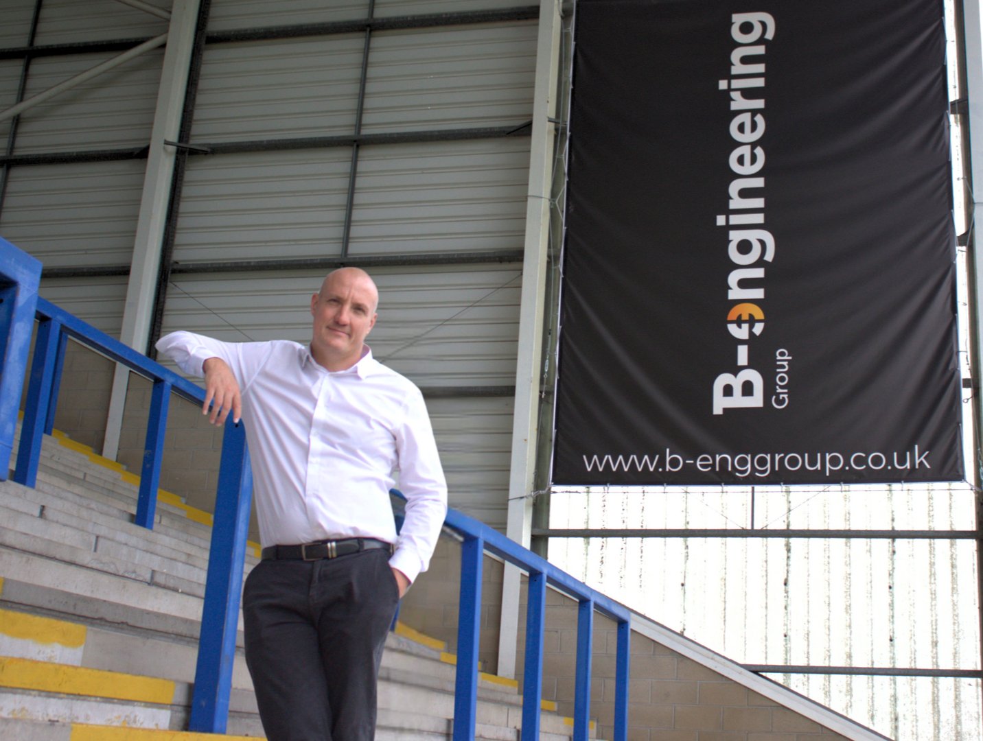 Man in white shirt leans on blue railing in industrial setting. Large banner reads "B-engineering Group" with URL. Neutral expression.