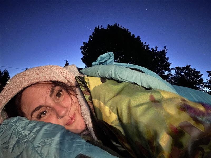 Person in a beanie lying in a sleeping bag under a clear, twilight sky with trees silhouetted. Cozy and serene mood.