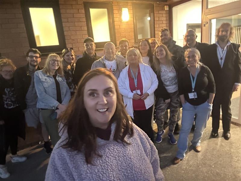 Smiling group of people pose outdoors at night, with a woman in a gray sweater in the foreground. Brick building and lit window behind.
