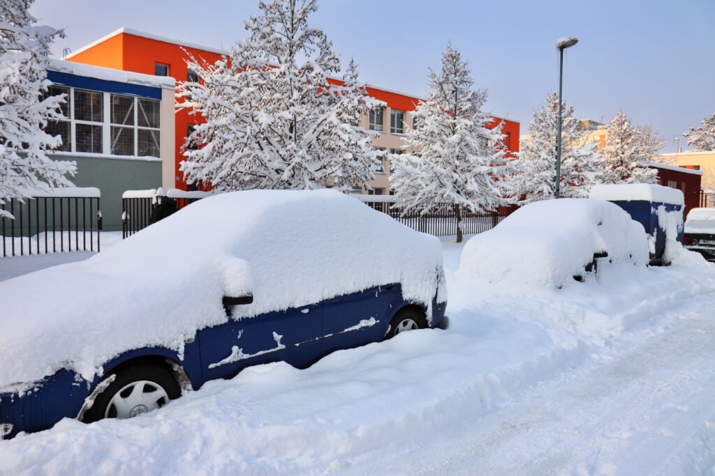 Snow-covered cars line a street by colorful buildings. Snow-laden trees stand nearby, creating a serene winter scene.