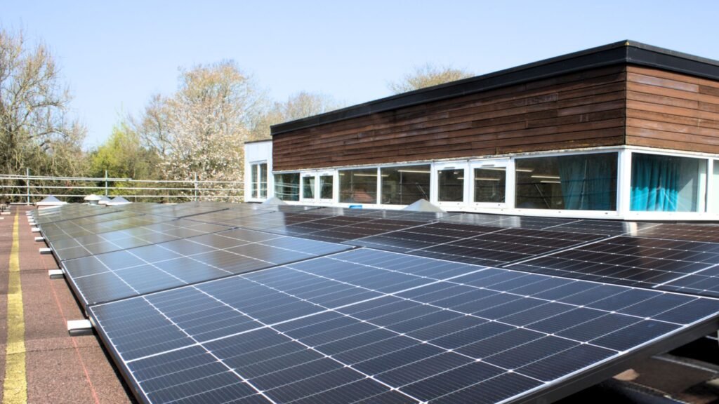 Solar panels on a rooftop reflect sunlight. Behind them is a building with wooden panels and windows. Trees are visible in the background.