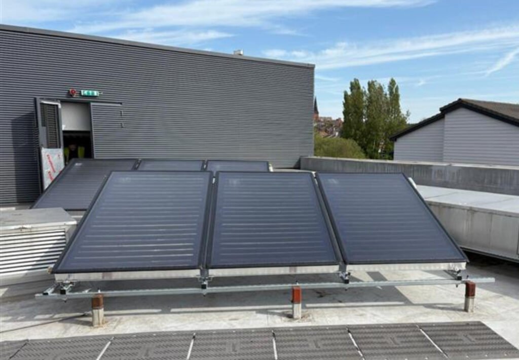 Solar thermal panels on a rooftop in front of a gray building. Open door with exit sign, trees in background, clear blue sky above.