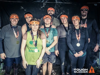 Team of 7 dressed in sports wear and covered in mud and dirt. they all have orange headbands with 15k tough mudder written on it. 
