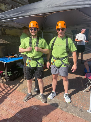 Two men in orange hard hats, green shirts and harnesses prepare for a challenge.