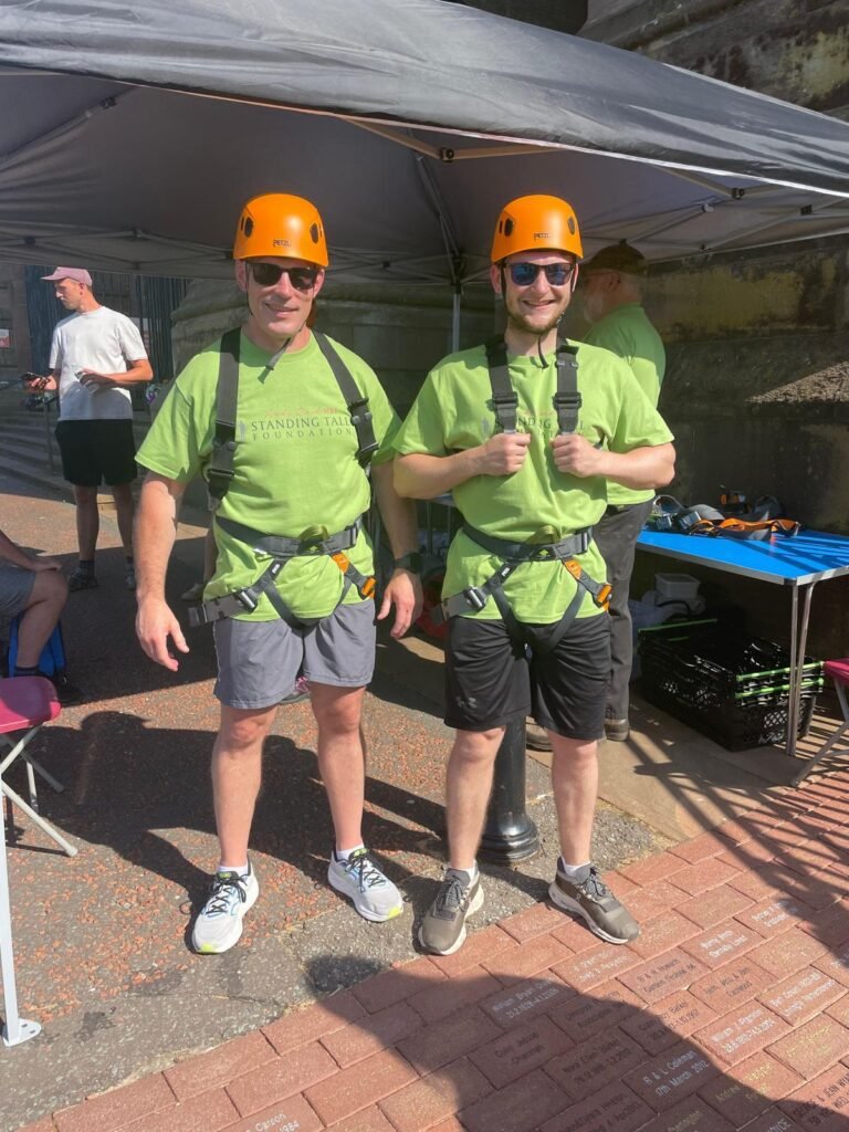 Two men stand in shorts and a green t-shirt and orange safety helments. Both are wearing a harnest.