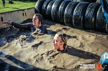 Two women are in muddy, cold water. They are shocked from the cold. A line of black tires are lined up behind them. 
