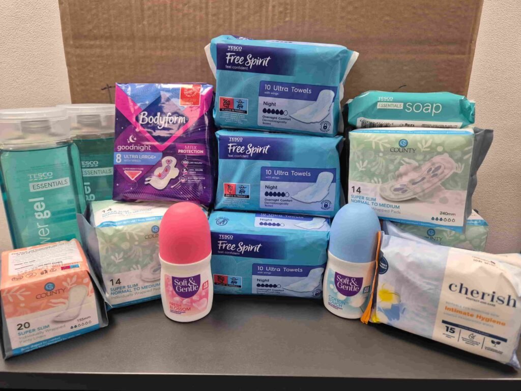Various personal hygiene products, including sanitary pads, soap, and deodorants, arranged on a table. Brands and packaging visible.