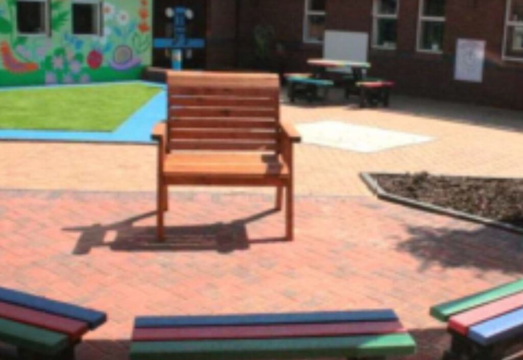 Wooden bench in a colorful schoolyard with mural, grass, and brick patterns. Surrounding is a circular seating area with multicolored benches.