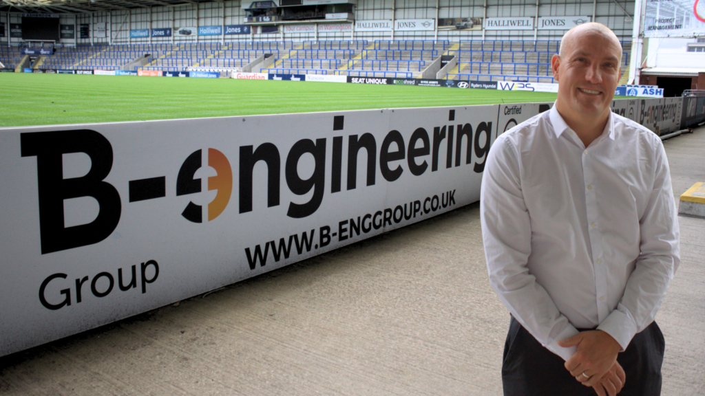 Man in a white shirt smiling by a stadium pitch. Engineering ad on barrier. Background: empty stands, mix of blue and grey seats.